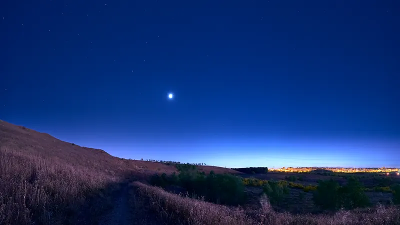 Ladera del Cerro Almodóvar, en Vicálvaro, al anochecer, con el cielo azul, un planeta brillante y las luces de Madrid en el horizonte