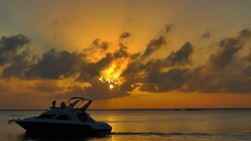 Sol escondido entre nubes sobre la Laguna Nichupté de Cancún, con un yate en silueta pasando frente a Plaza La Isla