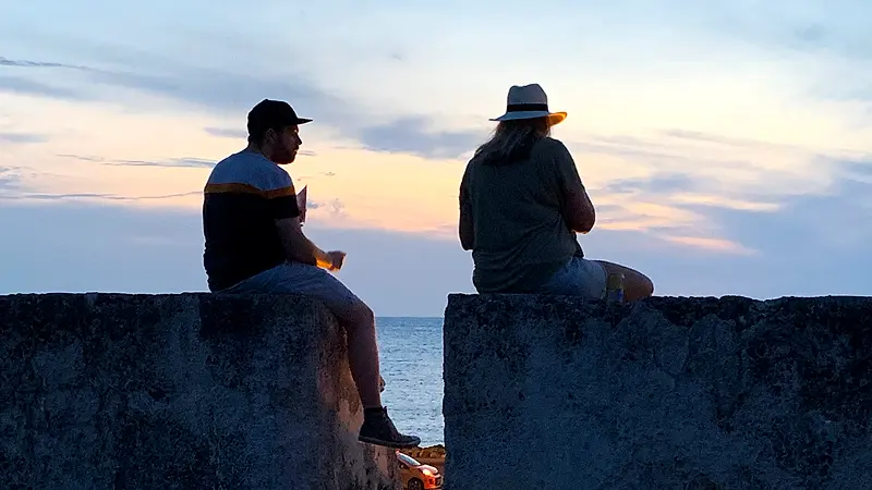 Dos personas sentadas en la muralla de Cartagena mirando el atardecer sobre el mar