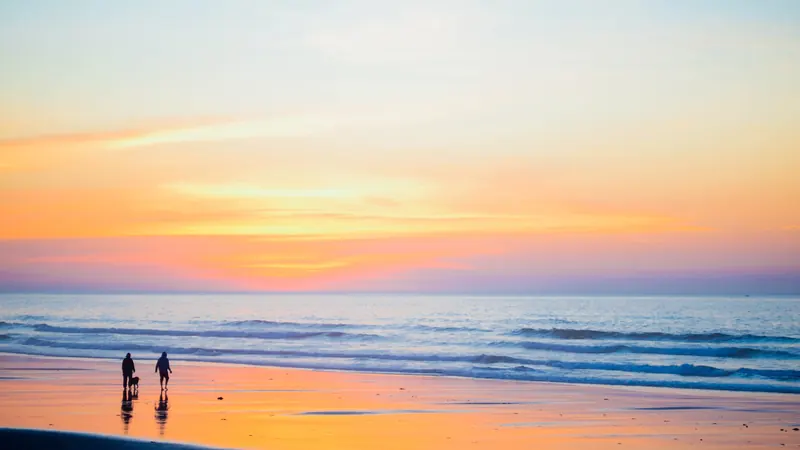 Dos personas pequeñas en la orilla de una playa mirando el cielo rosado y naranja del atardecer
