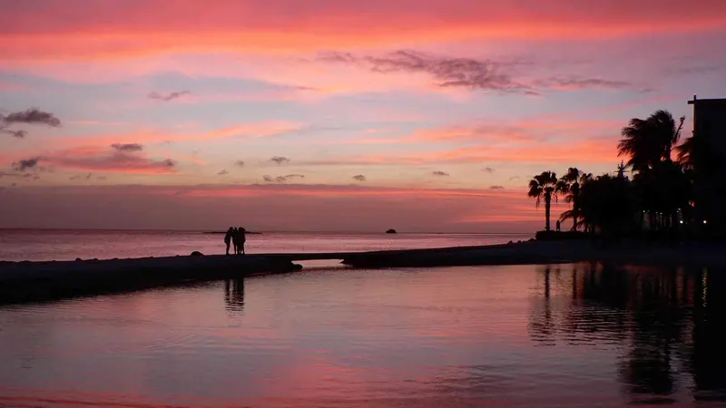 Dos personas en un espigón frente al mar Caribe en Oranjestad, Aruba, bajo un cielo rosa después de la puesta de sol