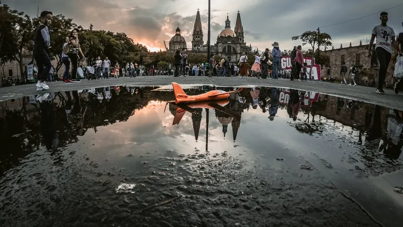 Gente paseando por la plaza junto a un charco que refleja el cielo del atardecer, con las torres de la Catedral de Guadalajara al fondo