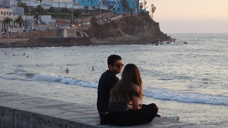 Pareja sentada en el muro del Malecón de Mazatlán con el mar y el cerro al fondo, al final de la tarde