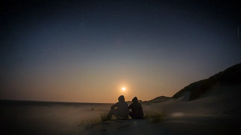 Pareja sentada en una duna mirando el último resplandor del sol en el horizonte, con el cielo ya estrellado