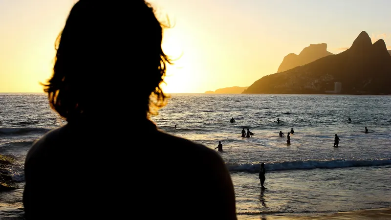 Silhueta de uma pessoa de costas vendo o pôr do sol na Praia do Arpoador, com o Morro Dois Irmãos ao fundo e banhistas no mar